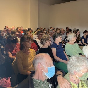 A large audience is seated closely together in a room, attentively watching an unseen presentation. The attendees are a mix of older and middle-aged adults, with some wearing face masks. The crowd fills the space, with people seated in rows, many facing forward with focused expressions. The lighting is soft, and the walls are plain, creating a neutral, indoor environment. The atmosphere appears to be one of concentration, as the audience listens quietly, likely during a lecture, presentation, or community meeting.