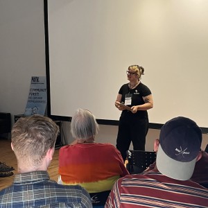 A person stands in front of a large white projection screen, speaking to an audience. They are wearing a black t-shirt, glasses, and a lanyard with a visible name tag, holding a booklet or tablet. There are three seated audience members visible from the back, two wearing striped shirts and one in a gradient red-to-yellow shirt. One person is wearing a navy baseball cap with the words "Big Dog" printed on it. To the left, a sign partially visible in the background reads "COMMUNITY FIRST! WE CAN END HOMELESSNESS," with the logo "PATH" at the top. The room has wooden floors and minimal decor.