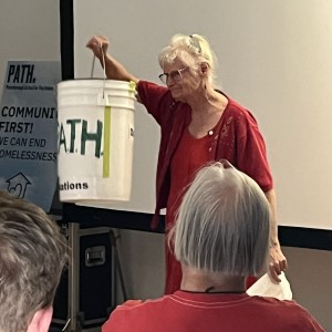 An older person with white hair, glasses, and wearing a red outfit, stands holding a white bucket labeled "PATH" in green letters. They are presenting to an audience, facing away from the camera, with one hand raised holding the bucket by its handle. The background includes part of the same "COMMUNITY FIRST! WE CAN END HOMELESSNESS" sign from the previous image. The audience member in the red-to-yellow gradient shirt is visible from behind, seated in the foreground. The setting is indoors, in front of a large projection screen.