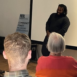 A person with dark hair in locks or braids, wearing a black outfit, stands in front of a seated audience, speaking with a relaxed posture and smiling. The audience members are facing away from the camera, including one person in a red-to-yellow gradient shirt. In the background, a partially visible sign reads "COMMUNITY FIRST! WE CAN END HOMELESSNESS," with the "PATH" logo at the top. The scene is indoors, with a large white projection screen and wooden floors visible. A couch and another seated individual are also partially visible on the left side of the image.