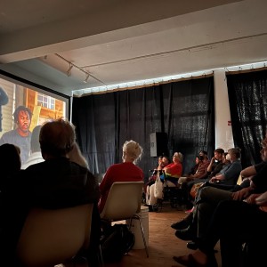 A group of people is seated in a darkened room watching a projected video. The screen shows a close-up of a person with locks or braids being filmed by a camera crew. The audience is seated in rows facing the screen, with several viewers visible in silhouette. A person in a red-to-yellow gradient shirt is sitting in the middle of the room, next to another person in a wheelchair. Black curtains line the walls, blocking out light, and there is a speaker and equipment set up near the back. The room has a casual, intimate atmosphere with wooden floors and simple white chairs.