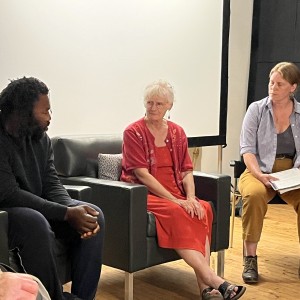 Three people are seated in chairs, engaged in a conversation. The person on the left, wearing a black outfit, sits with their hands clasped. In the middle, an older person with white hair and glasses, dressed in a red outfit, sits with their hands resting on their lap. The person on the right, wearing a striped shirt, mustard-colored pants, and boots, holds a notebook and appears to be listening attentively. They are seated in front of a large white screen in a room with wooden floors. The setting is casual, and the conversation seems part of a discussion or panel.