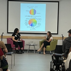 Two people are seated on stage in a conversation, facing an audience. One person, wearing a black shirt and seated in a pink chair, holds a microphone and a notebook. The other person, wearing a white top and black pants, is seated in a green chair, also holding a microphone. Behind them, a large screen displays an infographic with a colorful pie chart titled "What People Think PRIDE is" and "What PRIDE Actually is," showing comparisons between common misconceptions and the deeper meaning of pride. The audience is seated in rows, with some people visible from behind, including a person in a wheelchair and several wearing hats. A small table with water glasses is set between the speakers. The setting appears casual and educational, in a bright room with minimal decor.