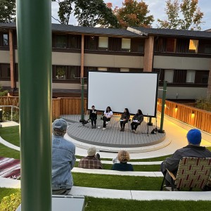 An outdoor panel discussion is taking place in a small amphitheater with tiered seating. Four people are seated on a circular wooden stage in front of a large white screen, engaged in conversation. The audience is seated on grass-covered steps, some on blankets or folding chairs. A few attendees wear hats or jackets, suggesting cooler weather. The setting is surrounded by a low wooden fence and modern multi-story buildings with large windows. Trees with autumn-colored leaves can be seen in the background, and soft lighting along the pathways adds warmth to the scene. The overall atmosphere is relaxed and open-air.