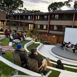 An outdoor amphitheater is filled with people seated on curved, grass-covered steps, attending a panel discussion. Four speakers sit on the small circular stage in front of a large white screen, engaged in conversation. The audience, wearing jackets and hats, is spread across the tiered seating, with some sitting on folding chairs or blankets. A striped blanket is laid out in one section, and several attendees are seated casually, while a few look at their phones. The scene is set against a backdrop of modern apartment buildings, trees, and soft evening light, giving the gathering a relaxed, community-focused atmosphere.