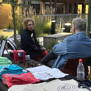 Two people sit at a merchandise table outdoors, conversing. One person, with curly hair and wearing a dark hoodie, faces the camera, while the other, with gray hair and a denim jacket, sits with their back turned. The table in front of them displays neatly folded t-shirts in bright colors (green, blue, and red), tote bags, and informational flyers. A plastic water bottle and other small items, like a cash box and a sign, are also on the table. The setting is in a landscaped courtyard with plants, illuminated by soft lighting from nearby fixtures and buildings in the background.