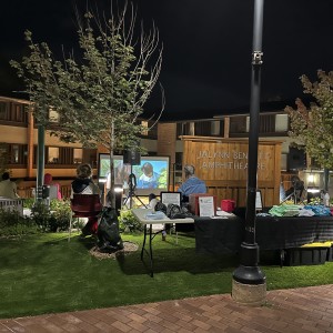 An outdoor event is taking place at night in the "Jalynn Bennett Amphitheatre," as indicated by a wooden sign on a building in the background. In the foreground, a merchandise table displays colorful folded t-shirts and other items under a streetlamp, with two people sitting nearby, one in a denim jacket and another in a hoodie. Behind them, a group of attendees watches a film projected onto a large screen. The amphitheater is landscaped with small trees, plants, and benches, softly lit by ground-level lighting. The atmosphere is calm, with people spaced out around the grassy area, engaging with the screening.