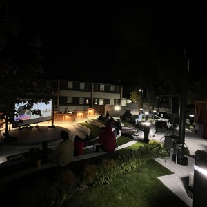 An outdoor film screening is being held at night in a small amphitheater. The large projection screen displays a movie scene, while the audience sits on tiered, grass-covered seating, spaced out and watching the film. Dim ground lighting illuminates the pathways and seating area, casting a soft glow over the steps and surrounding plants. Several people are visible sitting on benches or on the grass, with some blankets spread out. The surrounding buildings, including apartments with lit windows, form the backdrop, while the overall atmosphere is quiet and serene under the dark sky.