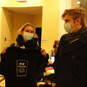 Two people, both wearing face masks, stand in an indoor setting. The person on the left, wearing a thick black scarf, holds up a black T-shirt with "ReFrame Film Festival" printed on it in white text. The person on the right, wearing a dark coat and striped scarf, looks at the shirt. In the background, a table with merchandise and a person seated behind it is visible, along with a small illuminated sign reading "ReFrame." The interior has soft yellow lighting, with red chairs and cream-colored walls, and a window reveals part of the street outside.