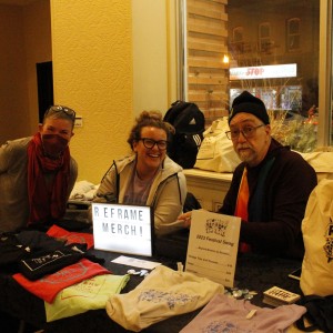 Three people are sitting at a merchandise table for the ReFrame Film Festival. The person on the left is leaning forward, wearing glasses, a red scarf, and a face mask, while the middle person, wearing glasses and a hoodie, is smiling. The person on the right is wearing a black beanie, glasses, and a dark-colored shirt, and sits in front of a rainbow-colored scarf. The table is covered with various festival-themed items, including T-shirts, tote bags, and buttons. A small illuminated sign on the table reads "ReFrame Merch!" and a printed sign next to it lists prices for "2023 Festival Swag," including T-shirts and tote bags. A large window behind them reveals the street outside, with the storefronts "First Stop" and "Liberty Tax" visible.