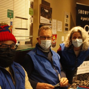 Three people, all wearing blue vests and face masks, are seated at a table in what appears to be a lobby area. The person on the left wears a red beanie with a colorful design, black glasses, and a thick cream-colored knit sweater. The person in the center has short, light hair, glasses, and a scarf, holding a pair of glasses in their hands. The person on the right has shoulder-length gray hair and leans slightly forward, smiling beneath their mask. Behind them, signage includes a poster reading "Showplace is your place," and various papers and posters are visible on the walls.