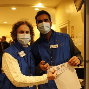 Two people are standing indoors, wearing blue vests and face masks. The person on the left has curly hair and is wearing a cream-colored knit sweater under their vest, with a name tag reading "Deborah." The person on the right has short dark hair and is wearing a name tag labeled "Abhi." They are holding a piece of paper together with a QR code printed on it, possibly related to event check-in or tickets. The background shows a softly lit interior with more people gathered, some also wearing masks, and informational signs on the walls.