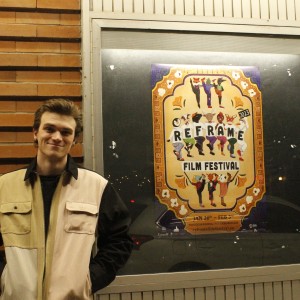 A person stands in front of a brick wall next to a large window displaying a colorful poster for the "ReFrame Film Festival." The individual is wearing a color-blocked jacket with beige, tan, and black sections, and they have their hands in their pockets, smiling slightly. The festival poster features stylized figures dancing in vibrant outfits, with the event name "ReFrame Film Festival" prominently displayed in bold letters. The event dates, "Jan 26th - Feb 3rd," and location, "Nogojiwanong/Peterborough," are listed below, along with a web address for more information.