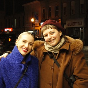 Two people are standing close together at night, smiling warmly at the camera. The person on the left is wearing a vibrant blue textured coat, has blonde hair pulled back, and is wearing red lipstick. The person on the right is dressed in a brown coat with a fur-lined hood, a burgundy beret, and a beige scarf, with short hair and a nose piercing. They are standing on a snow-covered sidewalk with parked cars and dimly lit buildings behind them. Signs in the background include "Liberty Tax" and other store signs, illuminated by street lamps.