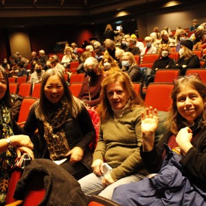 A group of four people sits together in a theater, smiling and interacting. The person on the left, wearing a black vest over a blue shirt with a colorful scarf, looks directly at the camera. Next to them , a person with long gray hair and a black vest smiles, holding a leopard-print scarf. The third person, wearing a green sweater, is seated, while the fourth person, holding a blue jacket, waves and smiles. Behind them, the theater is filled with people, many of whom are wearing face masks, seated in red chairs. A "RESERVED" sign is visible on a nearby seat. The setting is lively, with audience members preparing for or enjoying an event.