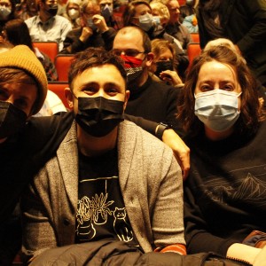 Three people sit closely together in a theater, all wearing face masks. The person on the left wears a mustard-yellow beanie and a black mask, leaning in toward the center. The person in the middle wears a gray cardigan, a black T-shirt with a design of plants and a cat, and a black mask. The person on the right wears a black sweatshirt with a mountain design and a light blue mask. Behind them, the theater is filled with people, many also wearing masks, seated in red chairs. The lighting is warm, highlighting the group in the foreground.