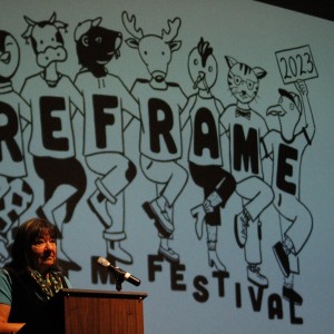 An older person with long, dark hair stands behind a wooden podium, looking forward. They are wearing a teal shirt layered under a black vest, along with a colorful scarf around their neck. A microphone is attached to the podium. In the background, a large projection displays cartoon animals, each wearing a shirt with a single letter, together spelling "REFRAME." One animal character holds a sign with "2023" written on it, and below the animals, the partial word "FESTIVAL" is visible. The stage lighting highlights the speaker against the playful, illustrated backdrop.