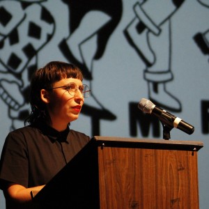 A person with short, dark hair, bangs, and round glasses stands behind a wooden podium, speaking into a microphone. They are wearing a black button-up shirt and have a focused expression. The background features part of the projection from the previous images, showing cartoon figures and partial text spelling out "FESTIVAL." The person is illuminated by stage lighting, standing out against the darker backdrop, with the microphone positioned in front of them on the podium.