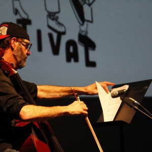 A musician is seated, playing a cello while adjusting sheet music on a stand in front of them. They are wearing a black hoodie and a backward baseball cap with a red underside, along with glasses. A tattoo is visible on their right forearm. The person is using a bow to play the cello, and their attention is focused on the sheet music. A microphone is positioned near the music stand, and part of the word "FESTIVAL" from the background projection is visible, though partially obscured by the musician. The lighting emphasizes the musician and their instrument against the dark stage.