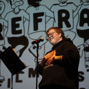 The same person stands at the microphone, now smiling with both hands placed over their chest. They are still wearing the black hoodie with the text "LOST A FRIEND TO AN OVERDOSE" and have visible tattoos on their forearms. The background projection remains the same, featuring cartoon figures and bold letters spelling "REFRAM" (likely part of "REFRAME FILM FESTIVAL"). The person’s posture and expression suggest an emotional or heartfelt moment, and the lighting continues to highlight them against the backdrop. A music stand with papers is positioned in front of the microphone.