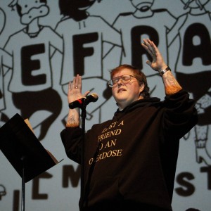 A person with short hair and glasses stands at a microphone, raising both hands mid-gesture. They are wearing a black hoodie with the text "LOST A FRIEND TO AN OVERDOSE" printed in bold white letters on the front. The person has visible tattoos on both forearms. In the background is a large projection featuring cartoon-like characters and bold, stylized letters spelling "REFRAM" (part of "REFRAME FILM FESTIVAL"), with the full word partially obscured by the speaker. The stage lighting casts a strong contrast, highlighting the speaker and the projection behind them.