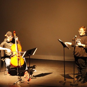 The two individuals on stage continue their performance, illuminated by focused lighting. On the left, the person playing the cello, seated and wearing a backward cap and dark clothing, is fully engaged with their instrument, bowing the strings. On the right, the person at the microphone appears to be singing or speaking passionately, mouth open wide in expression, with one hand gesturing outward. They are wearing glasses, a black hoodie with the phrase "LOST A FRIEND FOUND A CAUSE," and black boots. A music stand holds sheets in front of each performer. The stage remains bare, with the performers highlighted against a dark, minimalist backdrop.