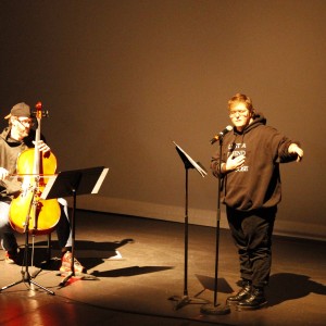 Two individuals are performing on a dimly lit stage. On the left, a person is seated playing the cello, wearing a black cap backward, a dark hoodie, and jeans. A music stand with sheet music is in front of them. On the right, another person stands at a microphone, mid-speech or performance, with one hand gesturing outward and the other resting on their chest. They are wearing glasses, a black hoodie with white text that reads, "LOST A FRIEND FOUND A CAUSE," and black boots. The stage is otherwise empty, and the lighting focuses on the performers, creating a dramatic contrast against the dark backdrop.