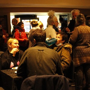 A group of people is gathered in a casual, social setting. Several individuals are seated at tables covered with dark tablecloths, eating and drinking. In the foreground, a person with blonde hair in a black sweater is smiling and chatting with someone whose back is facing the camera. Nearby, another person is drinking from a large blue water bottle, their face mask pulled down. In the background, more people are mingling, some wearing winter coats or scarves, and one person with long white hair tied in a high bun stands out. The room has soft lighting and a relaxed atmosphere, with many people holding plates of food or drinks.