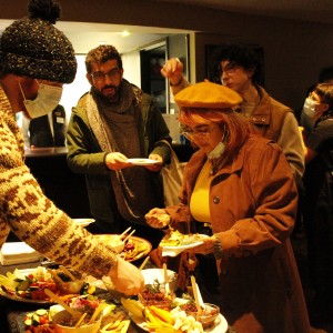 A group of people is gathered around a buffet table, serving themselves food. In the foreground, a person on the left, wearing a patterned knit sweater and a black beanie, is reaching for something on the table while wearing a face mask. On the right, a person with pink hair, glasses, and a mustard yellow beret, dressed in a brown coat, is adding food to their plate. Behind them, another person with a beard and glasses is holding a plate and appears to be waiting, dressed in a scarf and a green jacket. Others in the background are engaged in conversation, with a mix of casual and layered winter attire. The table is filled with various colorful dishes, including vegetables and small bowls of dip. The room has a warm, cozy atmosphere with soft lighting.