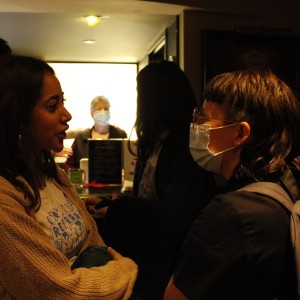 Two people are engaged in conversation in a dimly lit space. The person on the left, with long hair, is wearing a beige knit cardigan over a white T-shirt with blue graphic text and design. The person on the right, with a short mullet-style haircut and wearing glasses, is wearing a light blue face mask and a backpack. Both are facing each other, with a few others visible in the background, including someone wearing a face mask standing behind a counter, illuminated by brighter light. The environment suggests a casual gathering or event with a relaxed atmosphere.