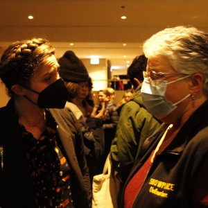 Two people are engaged in conversation in an indoor, crowded setting. The person on the left, with braided hair, is wearing a black face mask and a dark jacket over a patterned top. The person on the right, with short gray hair, glasses, and a light blue face mask, is dressed in a maroon shirt and a black vest with yellow embroidered text that reads "SHOWPLACE" and "Usher" on it. The background shows other people talking, some wearing winter clothing like beanies and jackets. The room is softly lit with recessed lights, creating a warm ambiance.