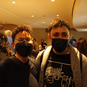 Two people are standing close together in a crowded indoor space, both wearing black face masks. The person on the left has short, curly hair, wears glasses, and is dressed in a dark top. The person on the right has short, tousled hair and is wearing a graphic shirt featuring line drawings of potted plants and a cat, under a light gray jacket. Behind them, other people are engaged in conversation or facing away, and the room is softly lit with recessed ceiling lights. The overall atmosphere suggests a social or casual event.