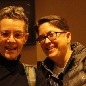 Two people are standing closely together, smiling at the camera. The person on the left has short, gray hair styled upward, wears light-colored glasses, a black turtleneck, and backpack straps over their shoulders. The person on the right has short dark hair, wears dark-rimmed glasses, and is dressed in a striped shirt with a puffy jacket. Both appear to be in a warmly lit indoor setting, likely at a social gathering or event. The background is softly blurred, with a dark monitor or screen visible behind them.