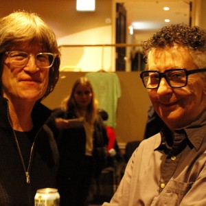 Two people are standing together, smiling at the camera in a warmly lit indoor setting. The person on the left has medium-length light brown hair, wears glasses and a black zip-up sweater, and is holding a can of drink. The person on the right has short, curly hair with shaved sides, wears large black-framed glasses and a gray button-up shirt, smiling with their arms crossed. In the background, a few other people are visible, including a person standing behind them holding a plate, and a wall with part of the words "THE NEXT" seen in the upper right corner. The setting suggests a casual social event.