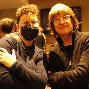 Two people are posing together, both crossing their arms and looking at the camera. The person on the left has short, curly hair, wears glasses and a black face mask, and is dressed in a dark sweater. The person on the right has medium-length light brown hair, wears glasses, and is dressed in a black zip-up sweater. Both have a casual, relaxed demeanor. In the background, other people can be seen mingling in a softly lit room, contributing to a warm, social atmosphere.