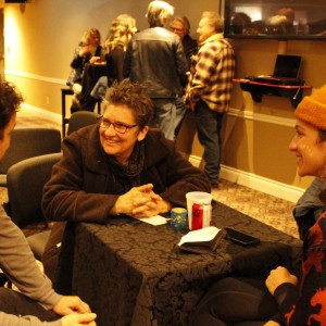 Three people are seated at a small table covered with a black tablecloth, engaged in conversation. The person on the left, wearing a gray long-sleeved shirt, is facing the other two. The person in the middle, with short hair, glasses, and a brown jacket, is smiling. The person on the right is wearing an orange beanie and a dark sweater with orange horse patterns. On the table are a can of Diet Coke, a white cup, and a phone. In the background, a group of people is standing and talking near a wall-mounted TV, while others are seated at taller tables, contributing to a casual, social gathering atmosphere.