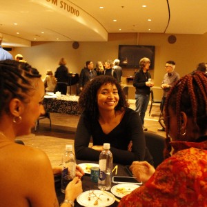 In the foreground, three people are seated at a table, engaged in conversation. The person in the center, with curly hair and wearing a black top, is smiling toward the camera. The two other individuals, seated with their backs to the camera, have braided hairstyles—one with a ponytail and hoop earrings, and the other wearing a red floral-patterned outfit. There are plates and water bottles on the table. In the background, more people are mingling and chatting in a softly lit room with a relaxed atmosphere. A black table with food is visible, and the curved ceiling has the word "STUDIO" partially visible. The gathering appears to be a social or networking event.