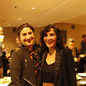 Two people are standing together in the foreground, smiling at the camera during what appears to be a social event. The person on the left has a braided crown hairstyle, wears a black jacket over a patterned blouse, and is smiling broadly. The person on the right has curly dark hair, wears a black top with a black jacket, and is also smiling. Behind them, several people are engaged in conversation or eating at a table with food. The room is softly lit, and the atmosphere is casual, with people mingling in the background.