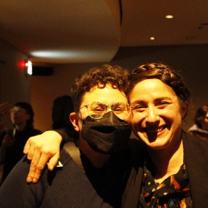 Two people are standing close together indoors, smiling for the camera. The person on the left has short curly hair, wears glasses and a black face mask, and is dressed in dark clothing. Their arm is around the person on the right. The person on the right, with a braided crown hairstyle, is smiling broadly and wearing a black jacket over a patterned shirt. In the background, several other people are engaged in conversation in the dimly lit room. The atmosphere suggests a social gathering or event.