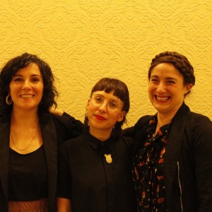 Three people are standing together, smiling at the camera against a light yellow wall with an intricate, embossed floral pattern. The person on the left has curly dark hair, wears hoop earrings, a black top, and a black jacket. The person in the middle has straight dark hair with blunt bangs, wears round glasses, a black button-up shirt, and a brooch shaped like an animal head. The person on the right has braided hair styled in a crown braid, wears a black jacket over a patterned shirt, and is smiling broadly. All three have their arms around each other.