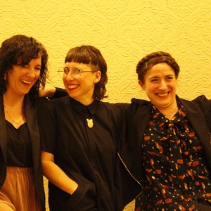 Three people are standing together, laughing and posing against a light yellow wall with an ornate floral pattern. The person on the left has curly dark hair, wears a black top with a black jacket, and beige pants, laughing with eyes closed. The person in the middle has straight dark hair with blunt bangs, wears glasses and a black button-up shirt with a brooch shaped like an animal head, smiling and looking to the left. The person on the right, with a braided crown hairstyle, wears a black jacket over a patterned blouse and has their arm outstretched, smiling broadly. They all appear to be having fun and sharing a light-hearted moment.