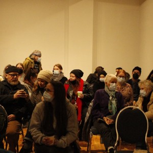 A group of people seated in a room, all wearing face masks. The individuals are dressed in warm clothing, such as jackets, beanies, and scarves, suggesting a cold environment. Some hold cups, and a few appear to be engaged in conversation while others look at their phones. The room has a plain light-colored wall with no visible decorations. The seating arrangement is informal, and the overall atmosphere is relaxed and social, possibly before the start of an event or lecture.