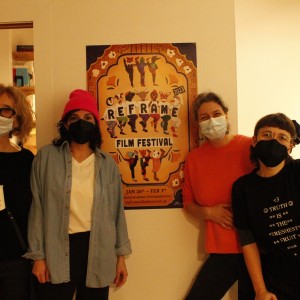 Four people stand together indoors in front of a "ReFrame Film Festival 2023" poster, all wearing face masks. The person on the far left has curly light hair, glasses, and holds a cup, wearing a black outfit. The second person, wearing a bright pink beanie, a light blue shirt, and a black mask, stands with hands in their pockets. The third person, in a bright orange sweater, smiles behind their mask. The person on the far right leans towards the center slightly, wearing glasses and a black shirt with white text that reads, "Truth is the freshest fruit." The background is simple, with some shelves visible in the adjacent room.