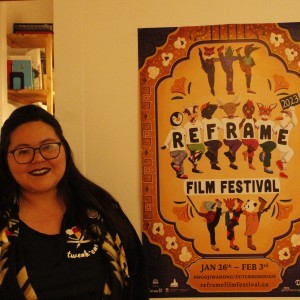 A person stands indoors, smiling, next to a colorful poster for the "ReFrame Film Festival 2023." The individual has long dark hair, glasses, and dark lipstick, wearing a black top with a design on it and a patterned cardigan. The poster features an illustrated group of people and animals holding up letters that spell "ReFrame," with vibrant patterns and figures dancing in various poses. The bottom of the poster lists the festival dates, "Jan 26th ~ Feb 3rd," and location, "Nogojiwanong/Peterborough," along with a website link and logos of sponsors. The background includes a shelf with books and a wall with a pink poster.