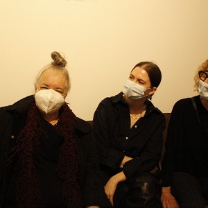 Three people sit together on a bench, all wearing face masks. The person on the left, an older individual with gray hair tied in a topknot, waves with their right hand and wears a dark coat and a thick scarf. The middle person, younger, with neatly pulled-back hair, looks toward the person on the left, wearing a dark buttoned shirt. The individual on the right, also older, has curly, light hair and wears large glasses, holding a disposable cup in one hand. The background is plain, with light walls and wooden flooring. The scene appears casual and relaxed.