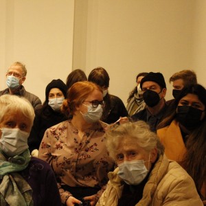 A group of people seated closely together in an indoor setting, all wearing face masks. The individuals vary in age, with some older adults in the foreground, and younger adults toward the back. Most of them are dressed warmly, wearing coats, scarves, and hats, suggesting a colder season. The background is plain, with light-colored walls, and a small sign visible on the right-hand side. The mood appears calm, possibly during a lecture or event.