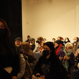 A crowded room is filled with people sitting and standing, all wearing face masks. In the foreground, a person with long hair, dressed in black, stands facing the camera. Behind them, a seated audience engages in conversation, some facing each other while others look toward the front. The group includes people of varying ages, with older individuals seated in the front rows. Many attendees wear warm clothing such as jackets, scarves, and hats. The room is dimly lit, with a dark curtain on the left and plain walls. The atmosphere is casual, with the audience appearing relaxed and engaged in pre-event or intermission conversations.