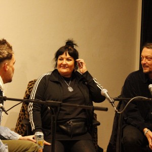 Three individuals are seated close together, engaged in conversation behind microphones, likely participating in a panel discussion. The person on the left wears a denim jacket over a white hoodie and holds a can of sparkling water or soda. In the center, a person wearing a black tracksuit with white stripes and a pendant necklace smiles, touching their ear. To the right, a person with longer hair, wearing a dark button-up shirt, listens attentively. The setting is casual, with relaxed body language and minimal background detail, focusing the scene on the interaction between the three. A chair with a leopard-print cover is partially visible behind the center person.