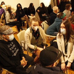 A group of people sit in conversation, wearing face masks in a warmly lit room with wooden floors. In the foreground, a person with long hair, a bandana, and glasses, wearing a black hoodie that reads "Lost a Friend to an Overdose," is seated with hands folded. Next to them, another person wearing a light cardigan and scarf engages in conversation with two others seated in front of them. To the right, a woman in a red sweater looks down at her phone. In the background, more people are seated, talking in small groups, with some gesturing as they speak. The overall atmosphere is casual and interactive, as attendees seem to be engaging in small discussions or waiting for an event to begin.