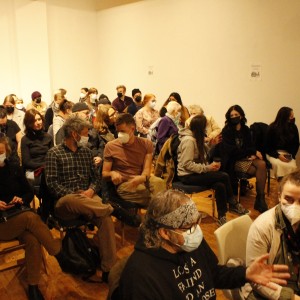 A masked audience sits in rows, facing forward in a well-lit room with wooden floors and plain walls. The crowd is diverse in age and attire, with most people wearing casual clothing such as sweaters, jackets, and scarves. Some individuals are seen engaging in conversations with those seated nearby, while others sit quietly or look at their phones. In the front, a person wearing a bandana and glasses gestures while talking to the person next to them. Several people hold coffee cups, and some are adjusting or resting in their chairs. The mood is generally relaxed, though the masks suggest an event occurring during a time of health precautions.